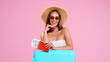 Positive Young Girl in straw hat and sunglasses With Tickets And Suitcase Pink Studio Background, smiles looking at camera
