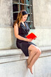 © Alexander Image - Sexy Woman Reading Outside. Wearing a black sleeveless trench coat dress, a hair band,  a young beautiful woman is sitting by a window, smiling, looking down, reading a red book.