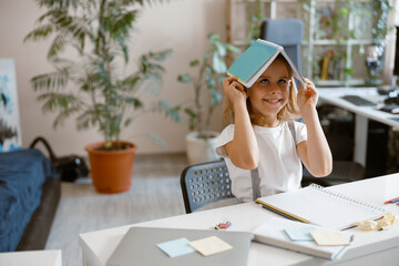 Wall Mural - Funny little schoolgirl holds copybook on head doing homework at home