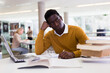 Thoughtful african-american man working with textbook and laptop in library. High quality photo