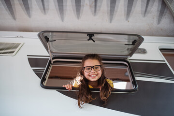 Wall Mural - Happy small girl looking out through caravan window and looking at camera, family holiday trip.