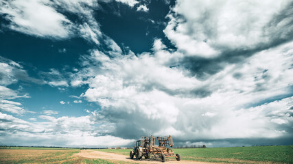 Wall Mural - Tractor With Attached Harrow In Sunny Spring Day. Countryside Rural Road Through Field Meadow Landscape Under Scenic Dramatic Sky With Clouds Before Rain