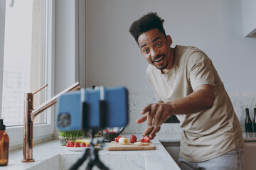 Wall Mural - Young surprised african american man in casual clothes prepare sweet breakfast cut fruit watch series on mobile cell phone cooking food in indoor kitchen at home alone Healthy diet lifestyle concept