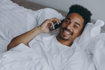 Wall Mural - Young cheerful happy african american man in nightwear talk on mobile cell phone lying in bed rest relax spend time in bedroom lounge home in own room house wake up dream be lost in reverie good day