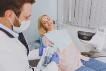 Young Man Stomatologist Doctor In Mask Open Disposable Sterile Tools Near Smiling Patient Caucasian Woman With Caries Sit At Dentist Office Chair Indoor Cabinet Healthcare Aesthetic Enamel Treatment.