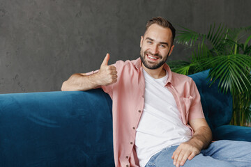 Wall Mural - Young smiling happy fun satisfied caucasian man 20s in casual clothes show thumb up gesture sitting on blue sofa at home flat indoors rest relax on weekends free time. People lounge lifestyle concept