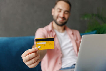 Canvas Print - Young man in casual clothes use work on laptop pc computer give close up focused credit bank card shopping online order delivery booking tour sitting on blue sofa at home flat indoors rest on weekend