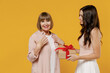 Side view two fun young surprised fun daughter mother together couple women in casual beige clothes gifting birthday present with red ribbon isolated on plain yellow color background studio portrait.