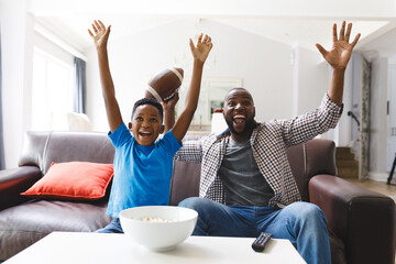 Wall Mural - Excited african american father and son sitting on sofa, watching match on tv and cheering