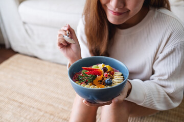 Wall Mural - A young woman holding and eating healthy blueberry smoothie bowl with mixed fruits topping at home