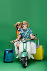 Wall Mural - cheerful middle aged and multiethnic couple in sun hats holding passports and luggage while riding moped on green