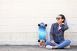 © interstid - Happy Asian woman wearing sunglasses with surfskate skateboard sitting against concrete wall