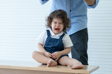 Little Caucasian kid girl with curly hair is sitting on wooden table crying. Her lips stained with tomato sauce and single dad standing behind at home