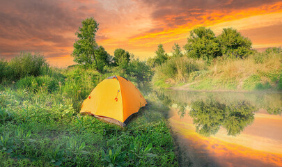 Wall Mural - orange tent on river bank in green grass under dramatic sky at sunset