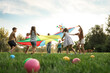 Group of children and teachers playing with rainbow playground parachute on green grass, low angle view. Summer camp activity