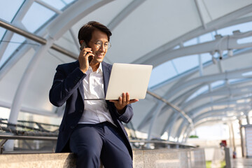 Asian confidently business man in glasses and suit working on laptop and mobole phone outdoor in city.