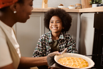 Cheerful little boy looking at his mom taking hot fresh apple pie out of electric oven in the kitchen