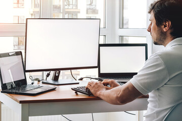 Wall Mural - Man sitting at workplace with two laptops and monitor near the window. Remote work from home