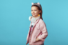 Stylish Little Girl In Pink Jacket And Kitty Headband, Holding White Headphones On Neck Looking At Camera Against Blue Background