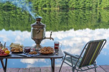 Wall Mural - Vintage metal tea samovar with white smoke and food on the table near the calm water lake in green forest at morning