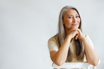 Wall Mural - Happy middle aged Asian woman with loose silver hair sits on chair leaning on hands on light background in studio. Mature beauty lifestyle