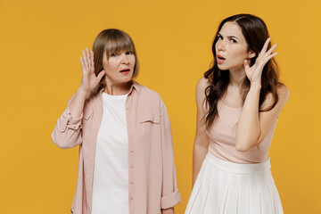 Two young curious nosy daughter and mother together couple women wearing casual beige clothes trying to hear you overhear listening intently isolated on plain yellow color background studio portrait