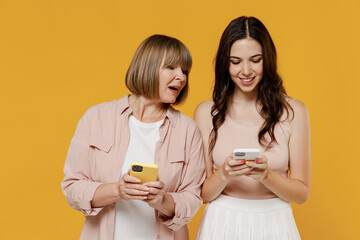 Two young smiling daughter mother together couple women in casual clothes hold in hand use mobile cell phone look at device peeps isolated on plain yellow background studio Family lifestyle concept