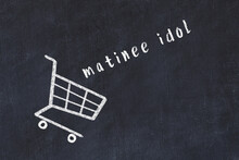 Chalk Drawing Of Shopping Cart And Word Matinee Idol On Black Chalboard. Concept Of Globalization And Mass Consuming