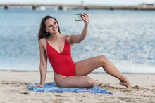 Woman In Red Swimsuit Sitting On Blue Mat On Beach Taking Selfie