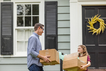 A father and daughter holding moving boxes outside a small blue cottage house getting ready for a move