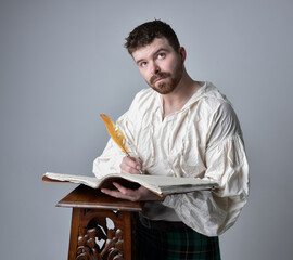 Close up portrait of handsome brunette man wearing Scottish kilt and renaissance white  pirate blouse shirt. Holding a quill and writing in a book,  pose isolated against studio background.   