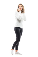 Wall Mural - Side view of young pretty woman in sweater wearing socks talking on the phone looking back. Full-length portrait isolated on white background