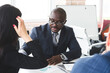 © xartproduction - Mature afroamerican businessman to discuss information with a younger colleague. People working and communicating while sitting at the office desk together with colleagues sitting.