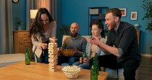 Group Of Friends Is Playing Game Of Building Tower Of Wooden Blocks In Evening, Woman Is Pulling Out Piece From Wobbly Building Squealing With Joy Jumping, Everyone Is Applauding And Enjoying Success