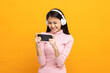 © Chanakon - Beautiful young asian women play mobile game and put on wireless headphone standing on isolated yellow background. Playing game on smartphone winning victory moment. Very enjoy and fun relax time