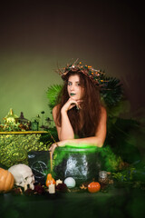 A witch with disheveled hair and long nails with a wreath of dry branches on her head, dressed in a green dress, brews a magic potion standing next to a cauldron and a table with ingredients.