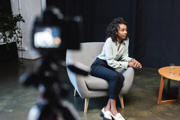 Wall Mural - brunette african american journalist sitting in armchair near coffee table and blurred digital camera