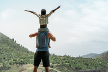 Back View Of Caucasian Father Holding On Shoulders Black Little Son On Top Of Mountain