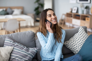 Wall Mural - Smiling woman on a sofa in a cozy interior and talking on a smartphone