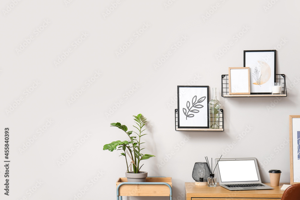 Modern workplace with laptop and frames near light wall