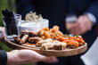 Delicious grilled bbq meet food platter with pork sausages dips and appetisers at wedding reception outdoors. Fresh tasty roast gourmet food on wooden board held by waitress.