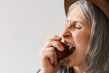 Wall Mural - Grey senior woman in hat eating apple with her eyes closed