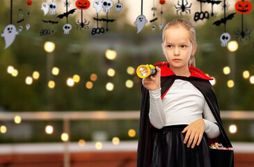 Wall Mural - halloween, holiday and trick-or-treating concept - girl in black dracula cape or costume with candies and flashlight over roof top party background