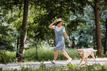 Wall Mural - young asian woman in sundress and straw hat running with dog in park