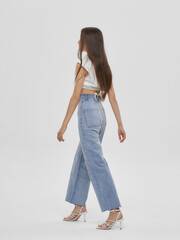 Wall Mural - Young elegant woman with long straight hair and natural make-up in white blouse and blue jeans standing and posing on bright grey background