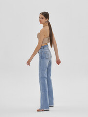 Wall Mural - Young elegant woman with long straight hair and natural make-up in beige top and blue jeans standing and posing on bright grey background