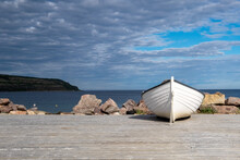 A White Wooden Fishing Boat On The Ocean Shoreline Under A Blue Sky With Lots Of Clouds.  A Land Point Is In The Background Meeting The Horizon. The Ocean Is Blue And Calm With Some Gulls On Rocks.