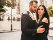 © halayalex - Beautiful fashion woman and her handsome elegant boyfriend in suit. Sexy brunette model in black evening dress. Fashionable couple posing in the street in Europe. Brutal man and his female outdoors