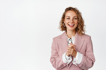 Young businesswoman clench hands, looking with hopeful face expression, smiling and looking at camera with anticipation, standing against white background