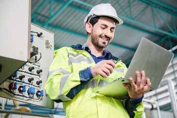 Young male machine inspector wearing vest and hardhat with headphones checking machine and sterilizers in water plant while making notes in digital tablet